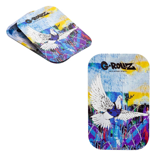 Two views of a G-Rollz Amsterdam magnetic rolling tray cover featuring a white dove with outstretched wings, blue and purple graffiti-style artwork, yellow accents, and black G-Rollz Amsterdam branding. One cover is shown flat, while the other is angled to show its thin, rounded rectangular shape.