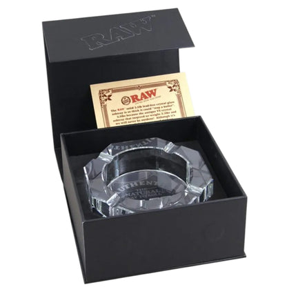 A clear glass RAW-branded ashtray displayed inside an open black presentation box with a branded insert card, on a white background. The Wee Smoke Shop