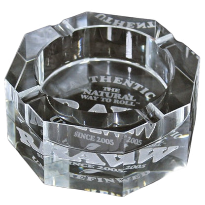 Clear faceted RAW glass ashtray with thick angled sides, built-in cigarette rests, and white “RAW,” “Authentic,” “The Natural Way to Roll,” and “Since 2005” branding visible through the glass. The Wee Smoke Shop